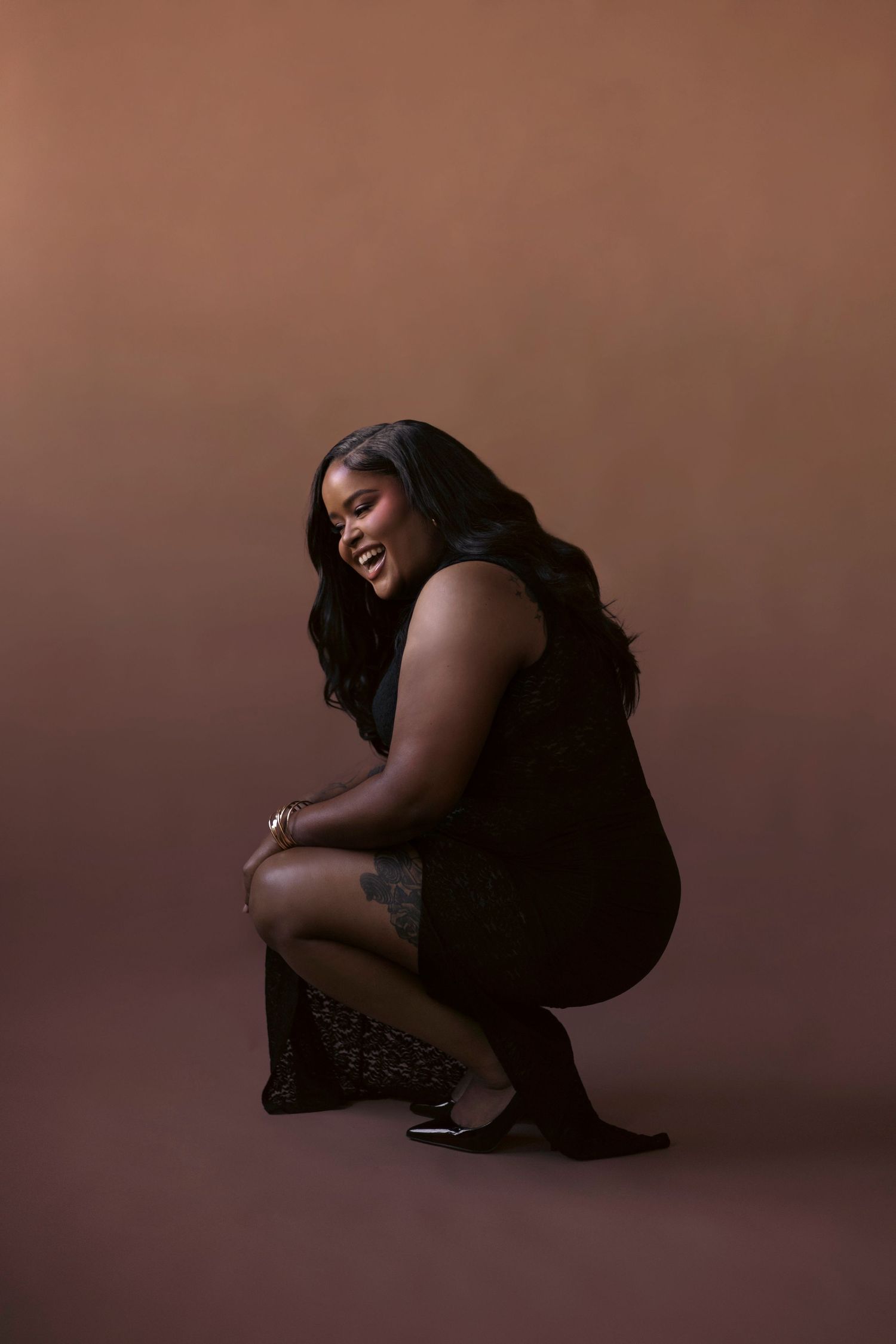 A silhouetted figure in black clothing crouches expressively against a dark brown studio backdrop.