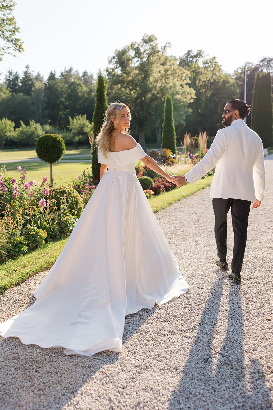 Newlywed couple walks down a gravel path surrounded by gardens and greenery on a sunny summer day.