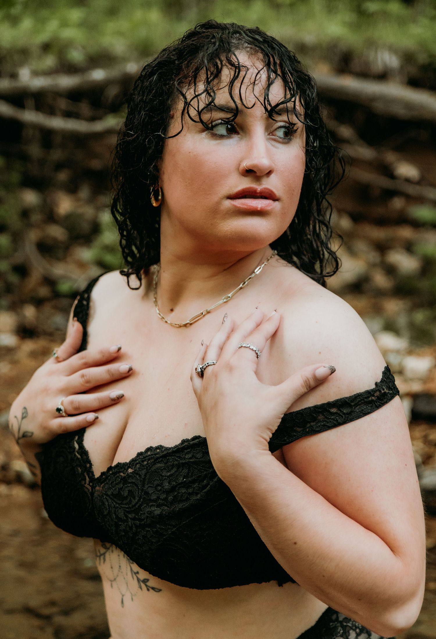 Portrait of a person with wet curly dark hair and a black lace top posing outdoors in a natural setting.