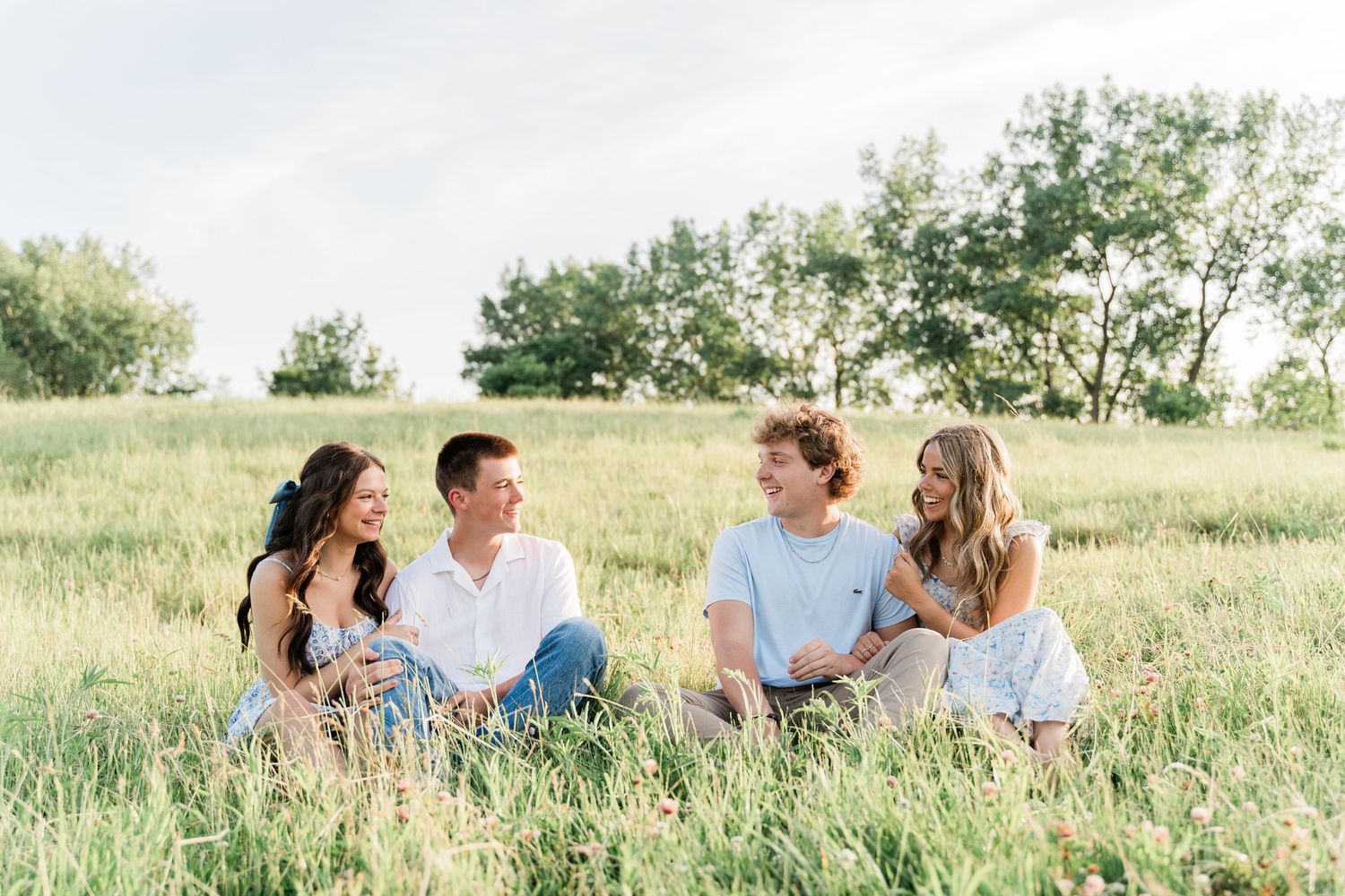 Group of people sitting and laughing together in a sunny green meadow during a casual outdoor photoshoot.