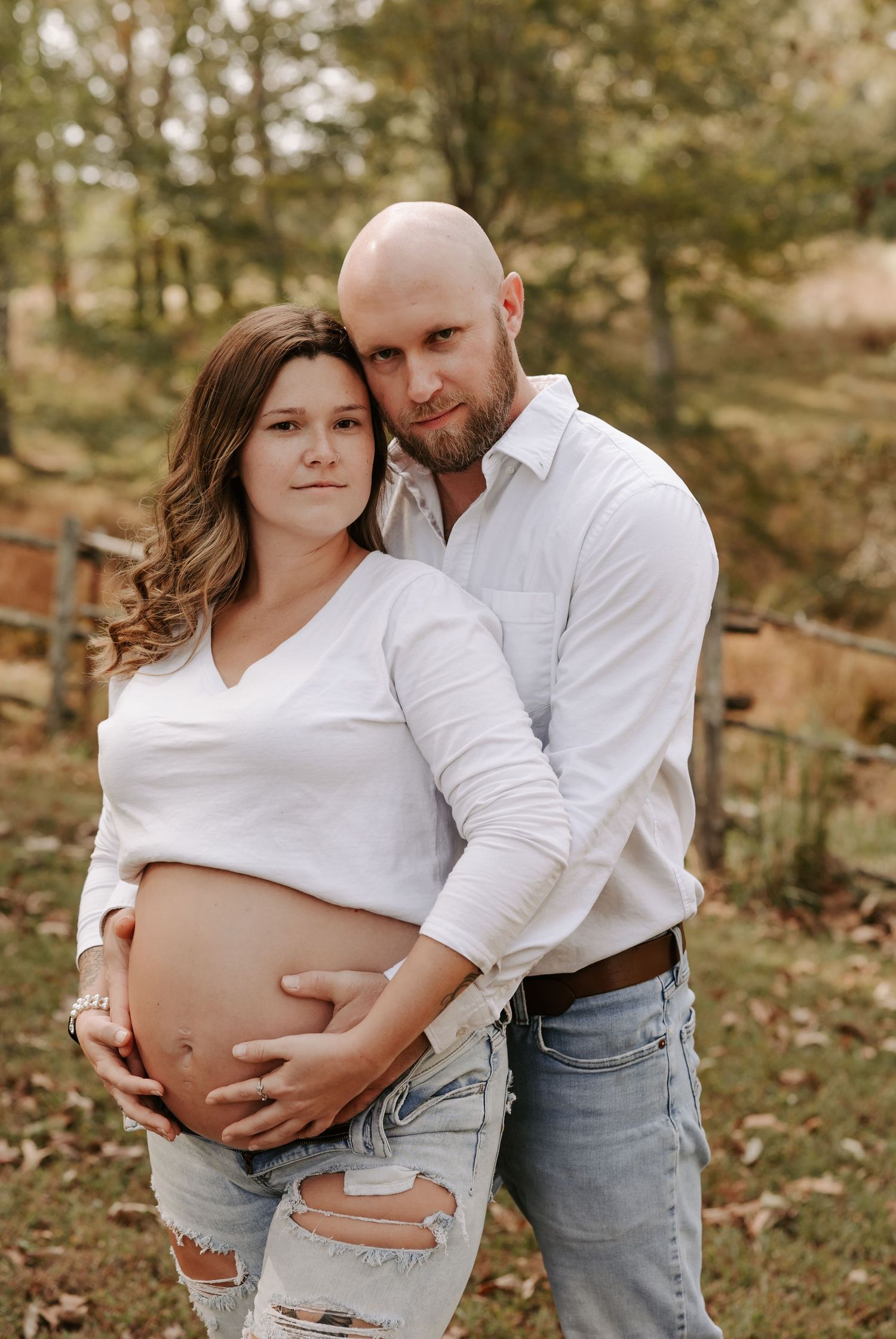 An autumn maternity photo featuring a couple in casual attire against a wooded backdrop.
