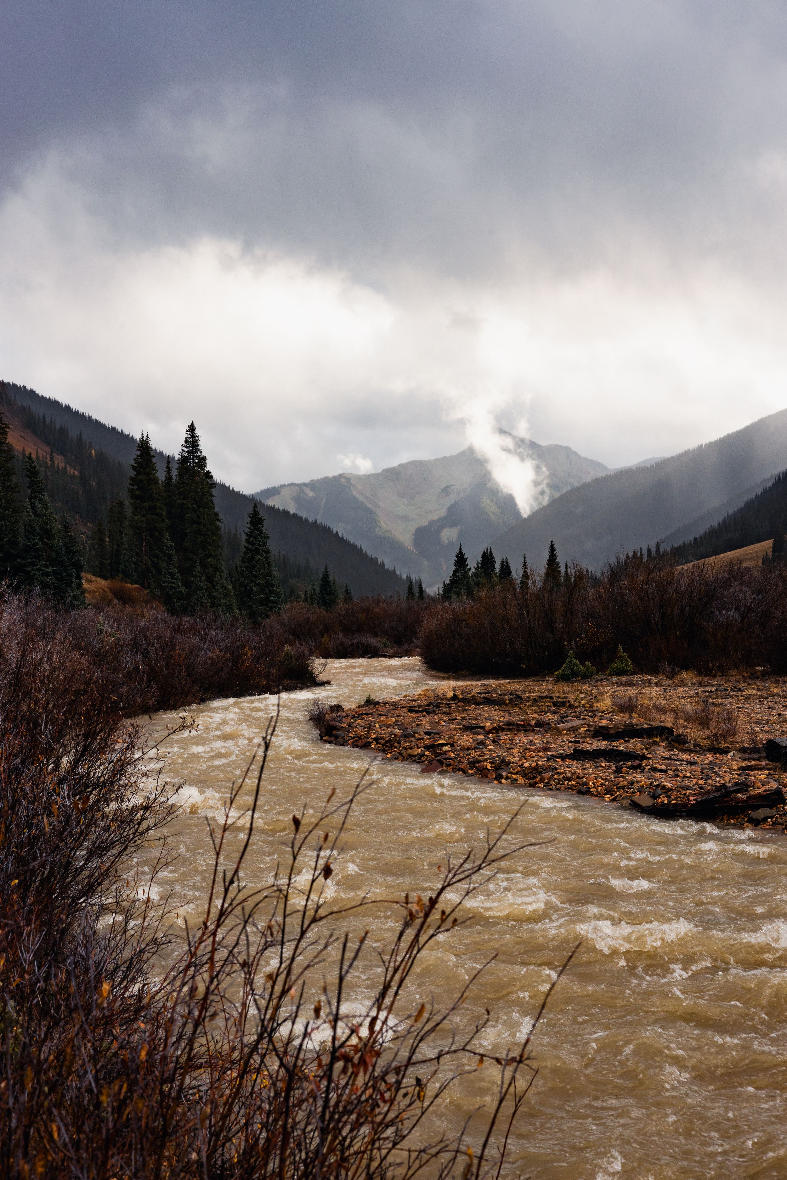 Dramatic mountain landscape with stormy clouds and rushing brown river through rugged terrain in autumn.