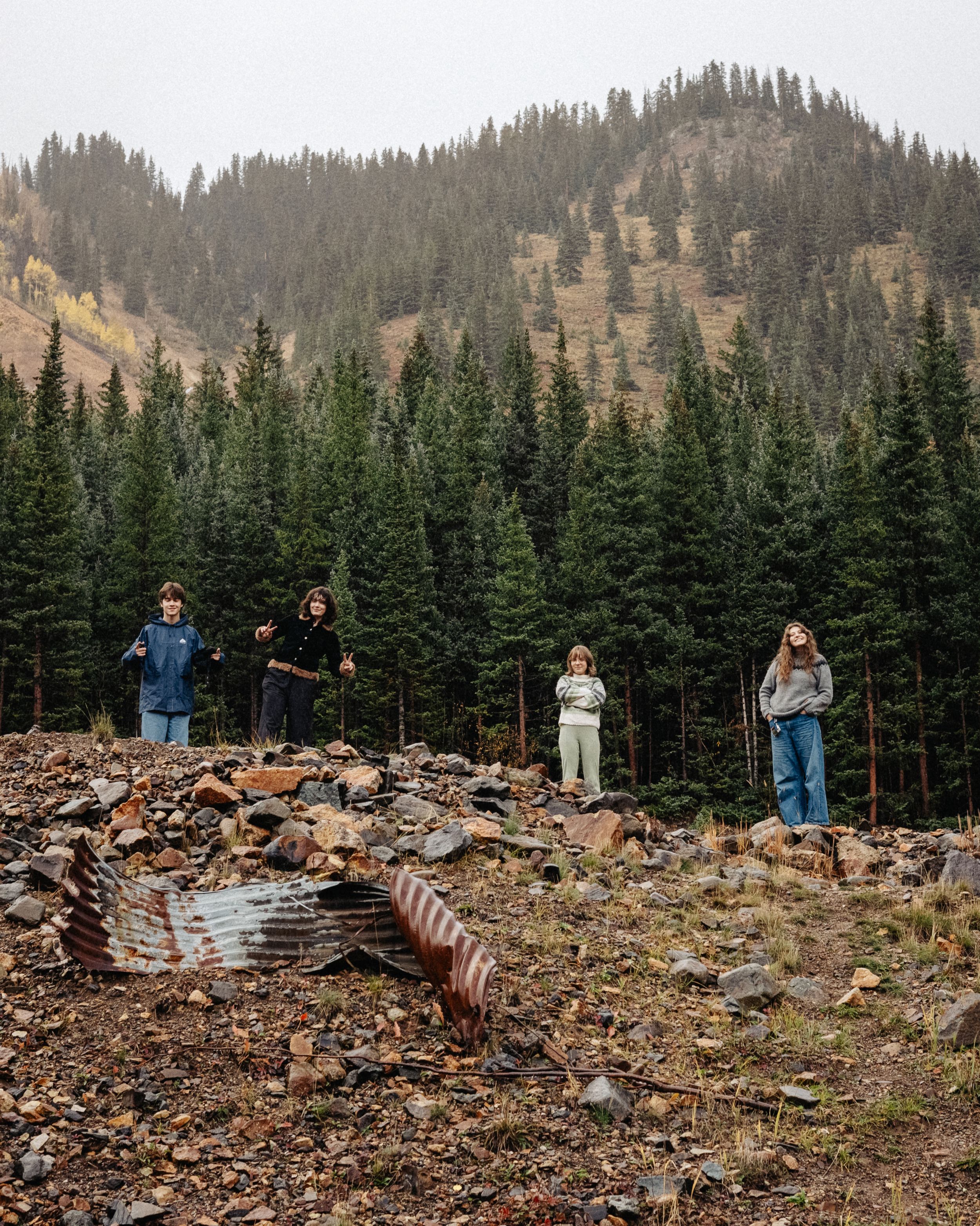Group stands on rocky mountain terrain surrounded by tall evergreen trees in a scenic wilderness landscape.