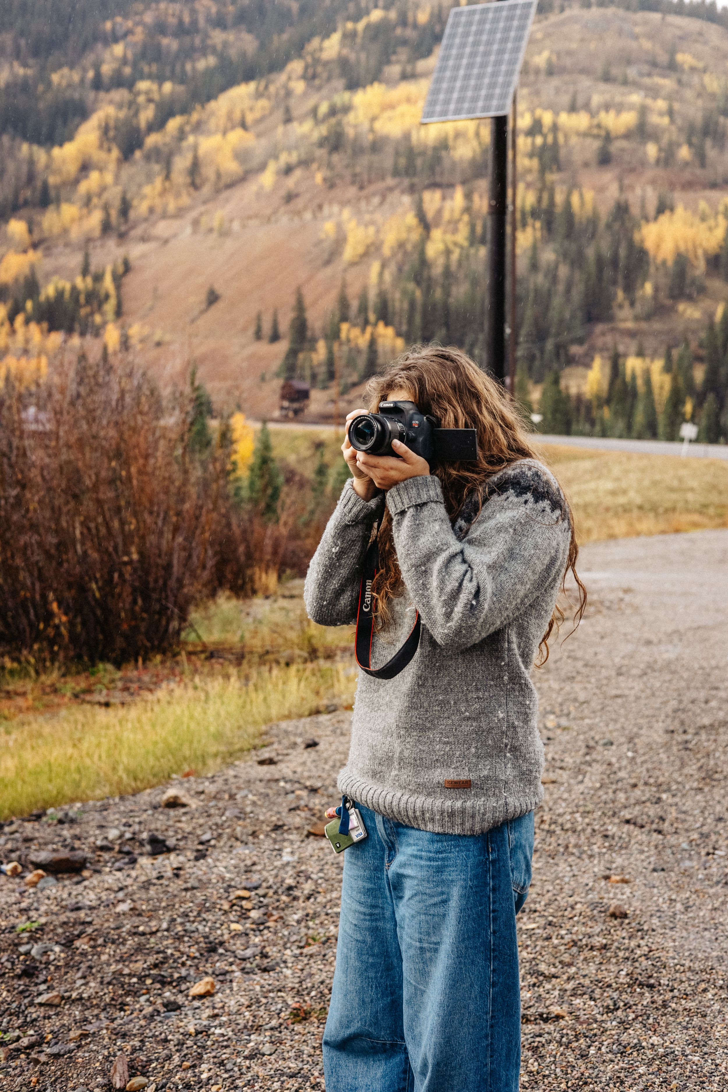 A photographer in a gray sweater and jeans takes photos against an autumn mountain landscape with golden foliage.