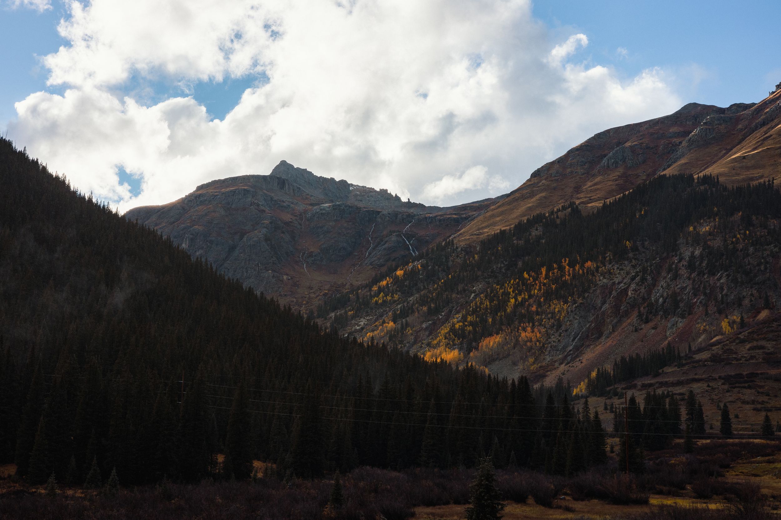 Panoramic mountain landscape with snow-capped peaks and dramatic clouds against a bright blue sky.