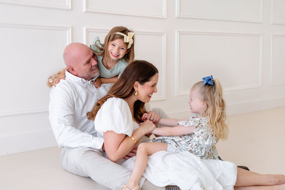 A family in white outfits sits together on the floor having a tender moment in a bright minimalist room.