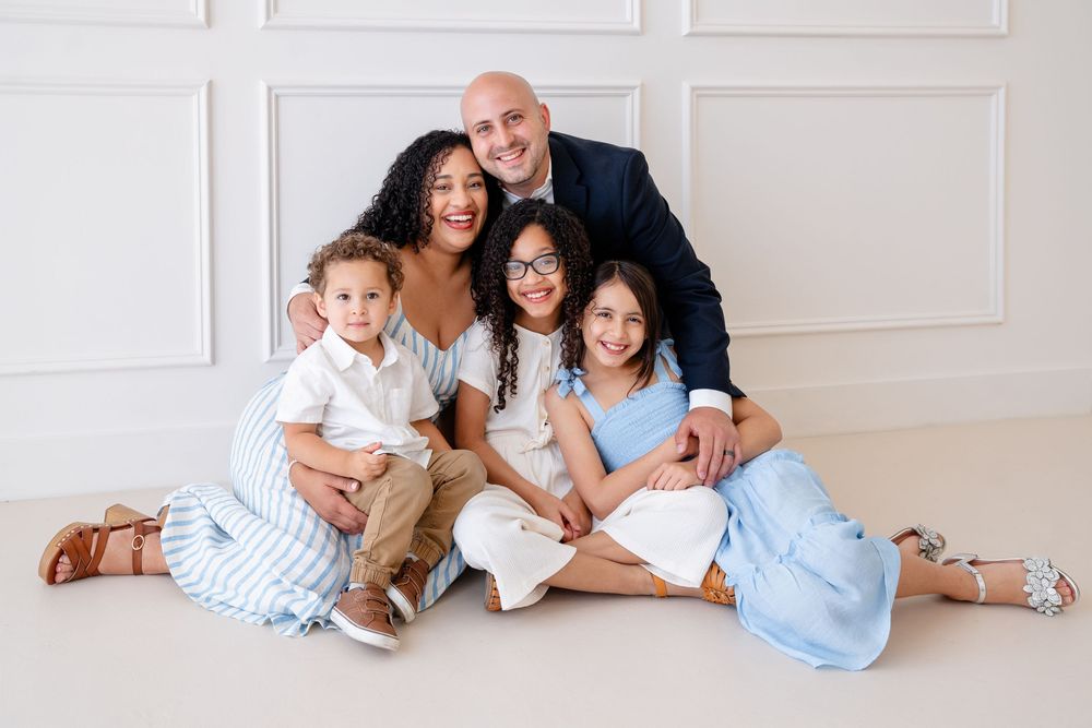 A family wearing coordinated white and blue outfits sits together on the floor for a portrait session.