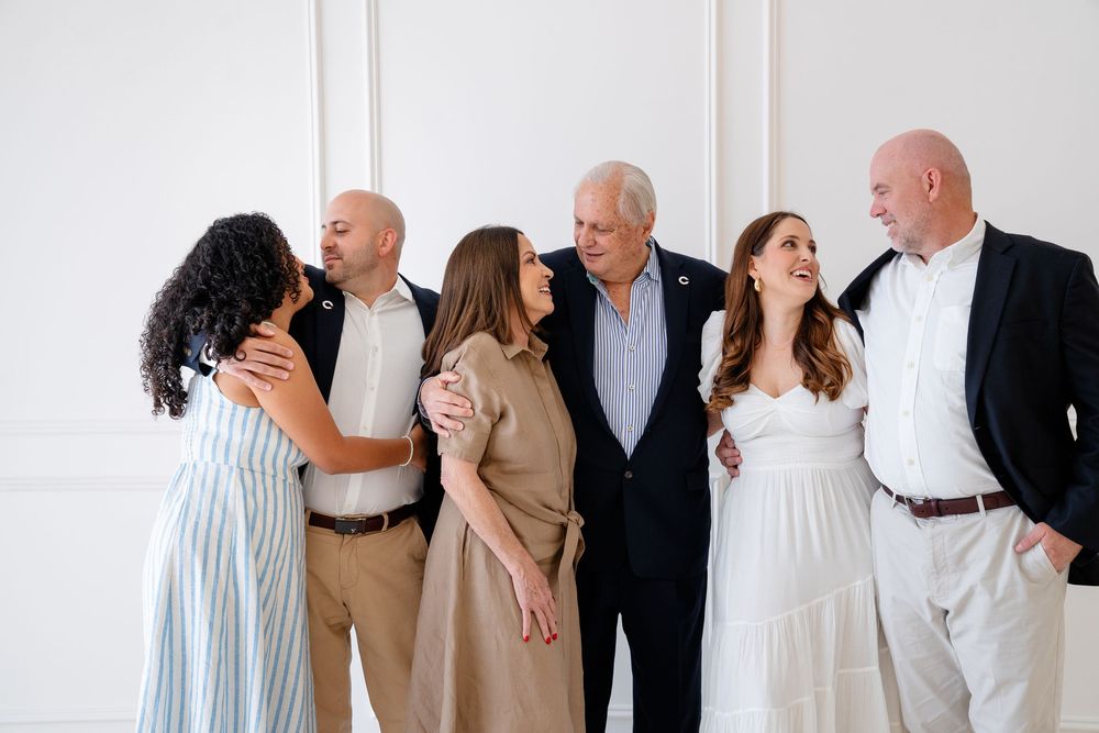 Multi-generational family group poses together in coordinated white and neutral outfits against white wall.