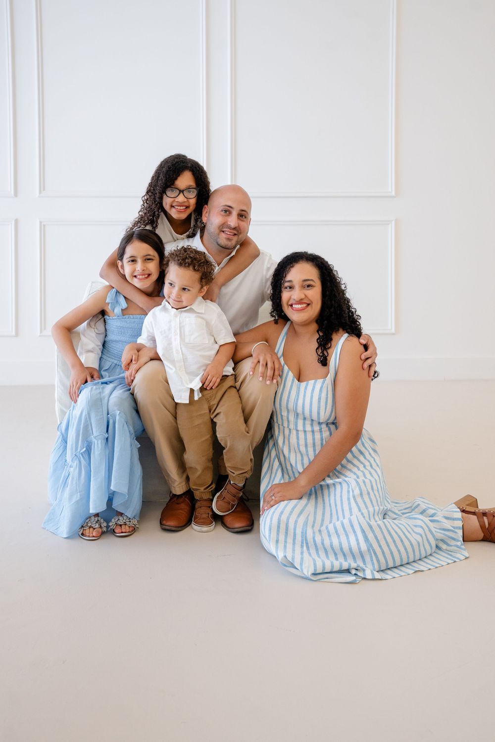A family in coordinated blue and beige outfits sits together on a white floor against a neutral wall for portraits.
