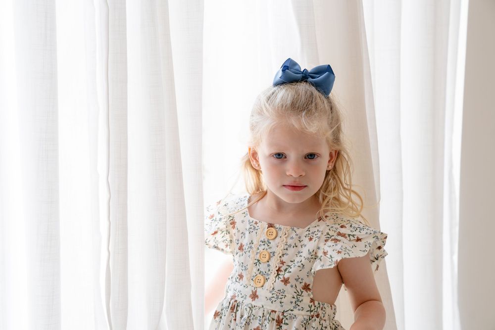 Young girl in floral dress stands against white curtain backdrop with natural lighting.