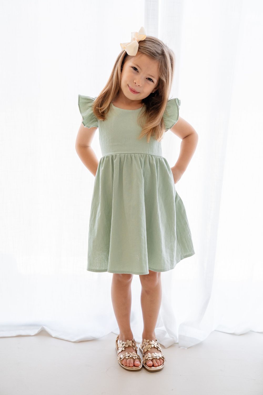 Child in mint green dress with ruffled sleeves and leopard print sandals poses against white background.