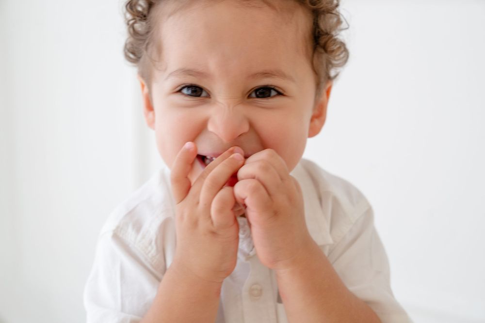 A happy baby in a white onesie smiles and covers their mouth with both hands in a playful expression.