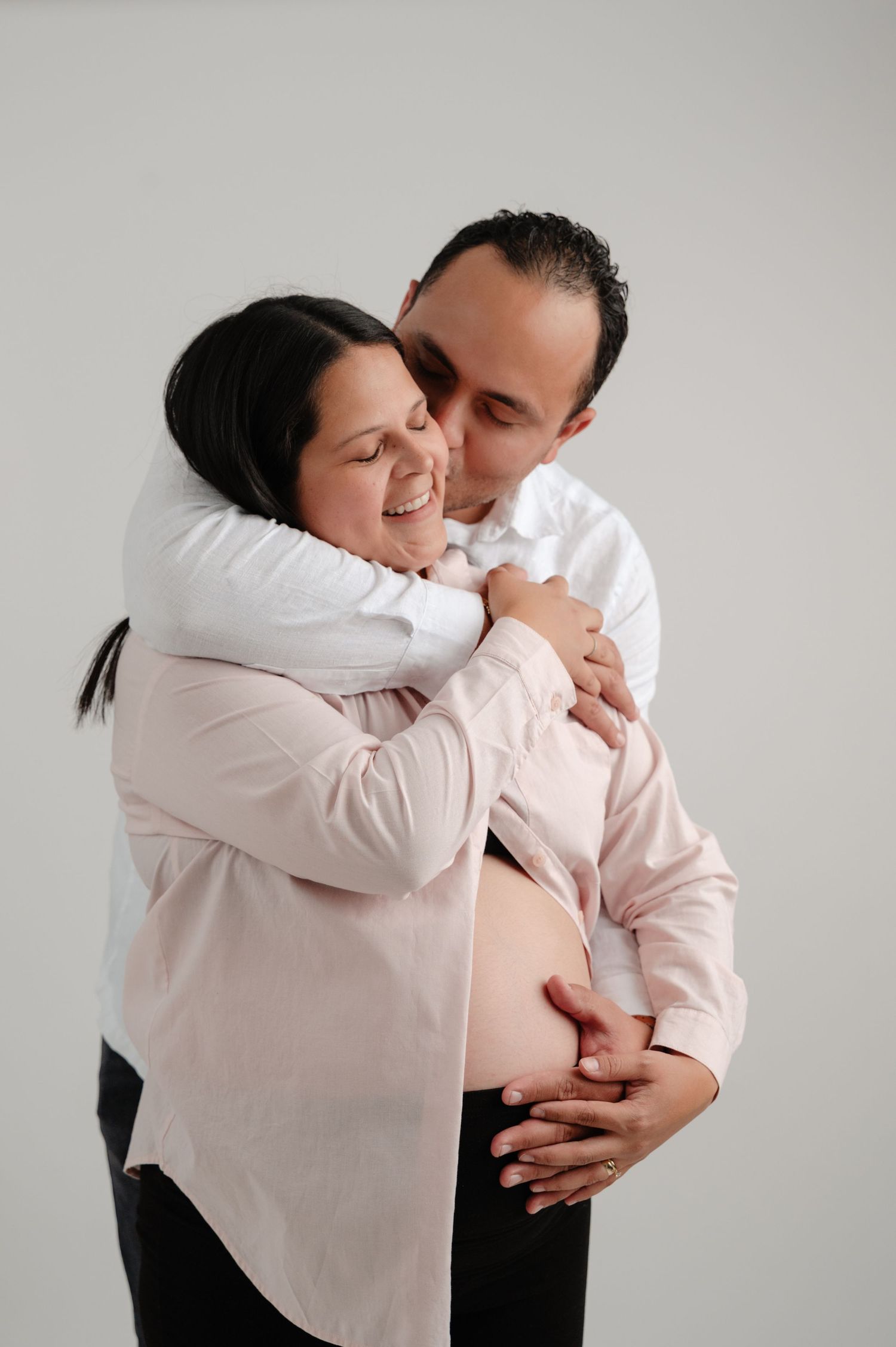 A couple wearing white and pink clothing embrace tenderly in a series of intimate maternity photos.