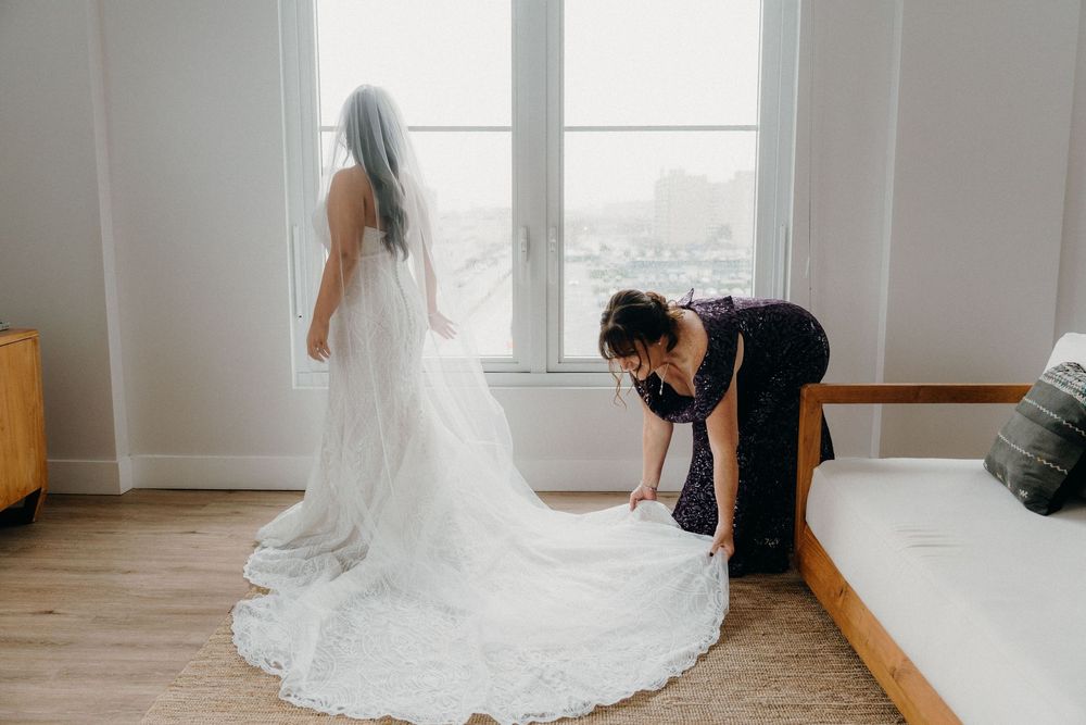 A wedding dress is adjusted and styled in a bright room with hardwood floors and large windows.