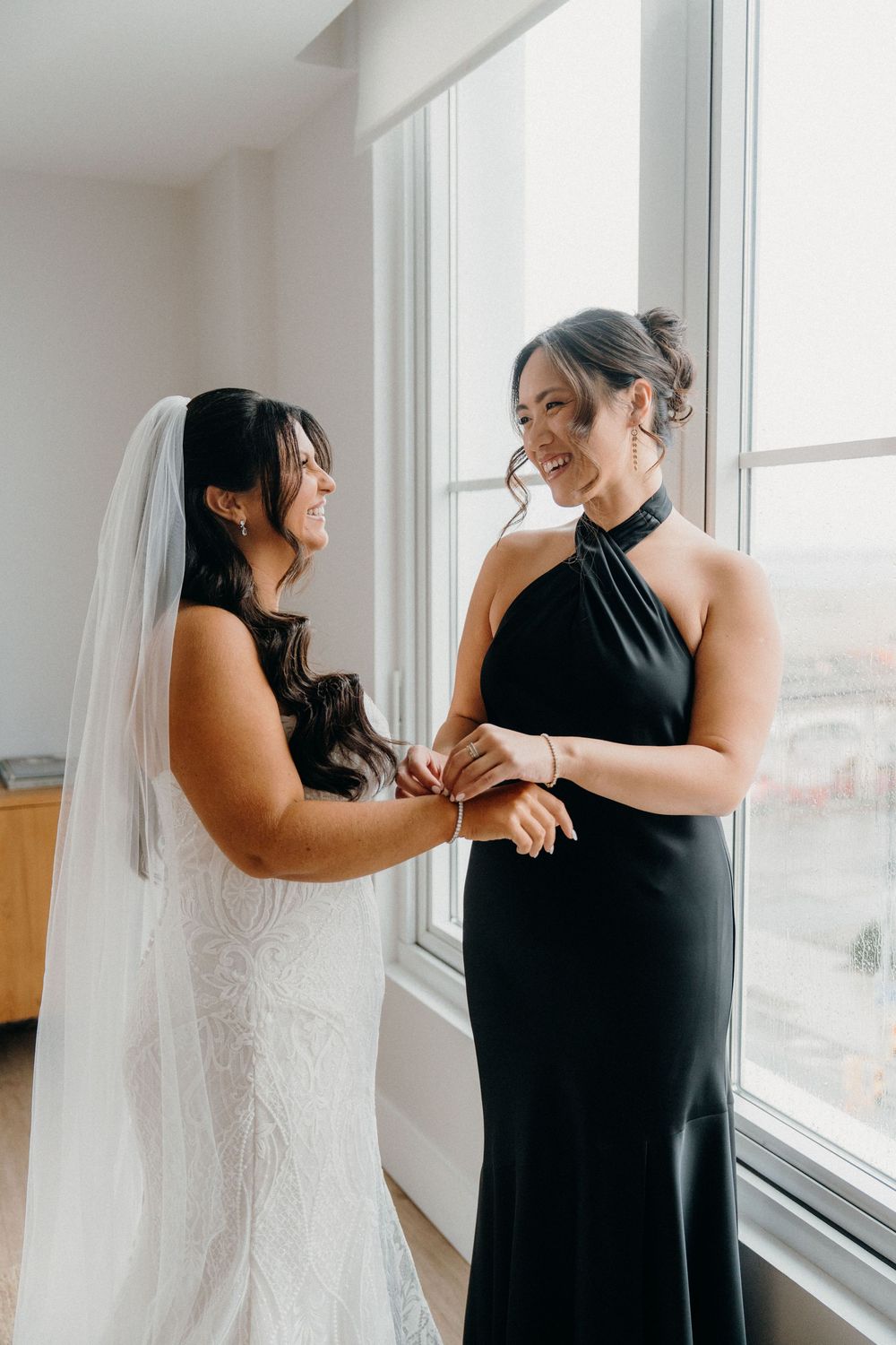 A touching moment captured in a bright room as someone helps adjust a white wedding dress near large windows.