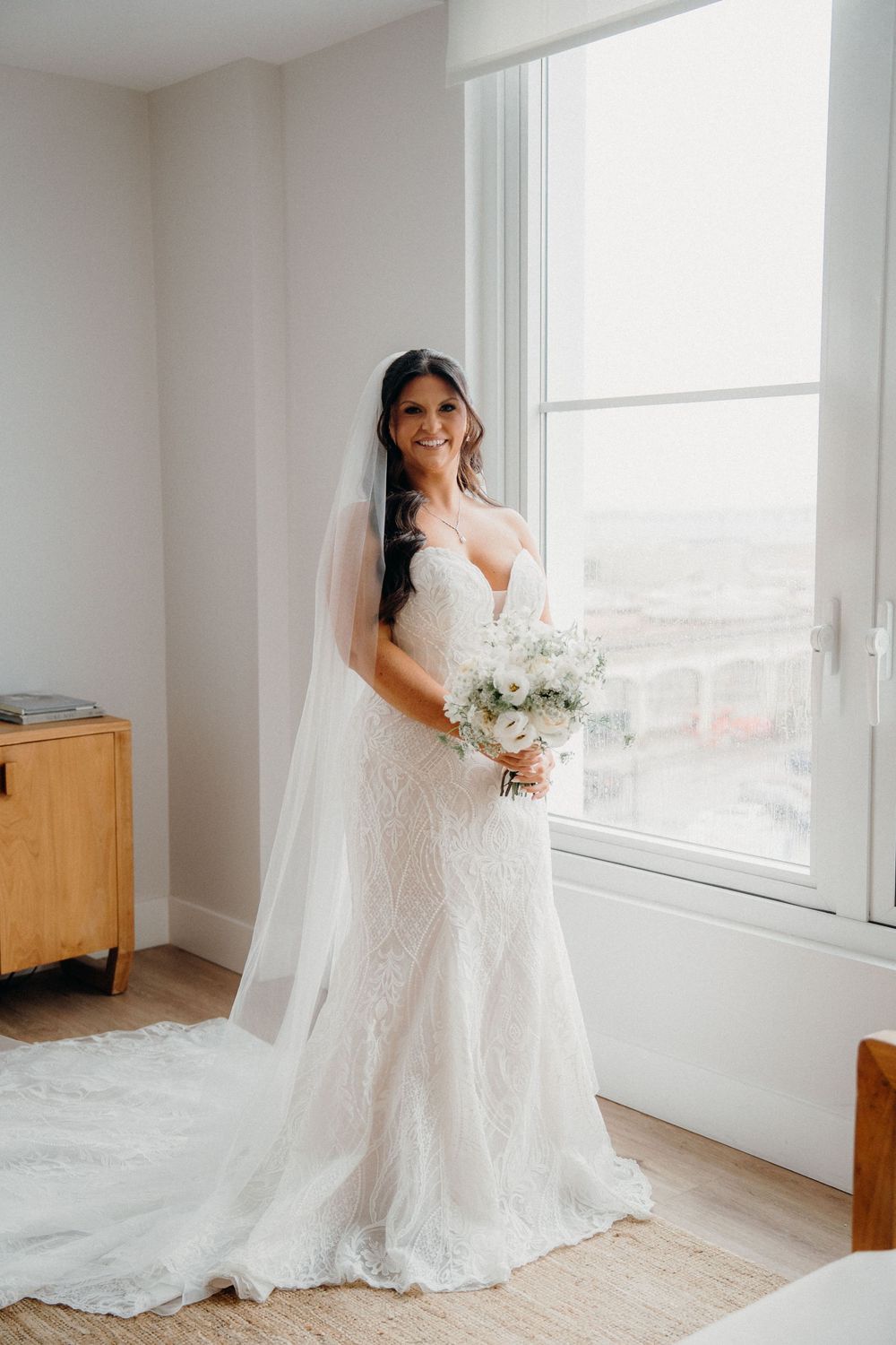 A bride in a lace wedding gown and flowing veil poses gracefully by bright windows in a minimalist white room.