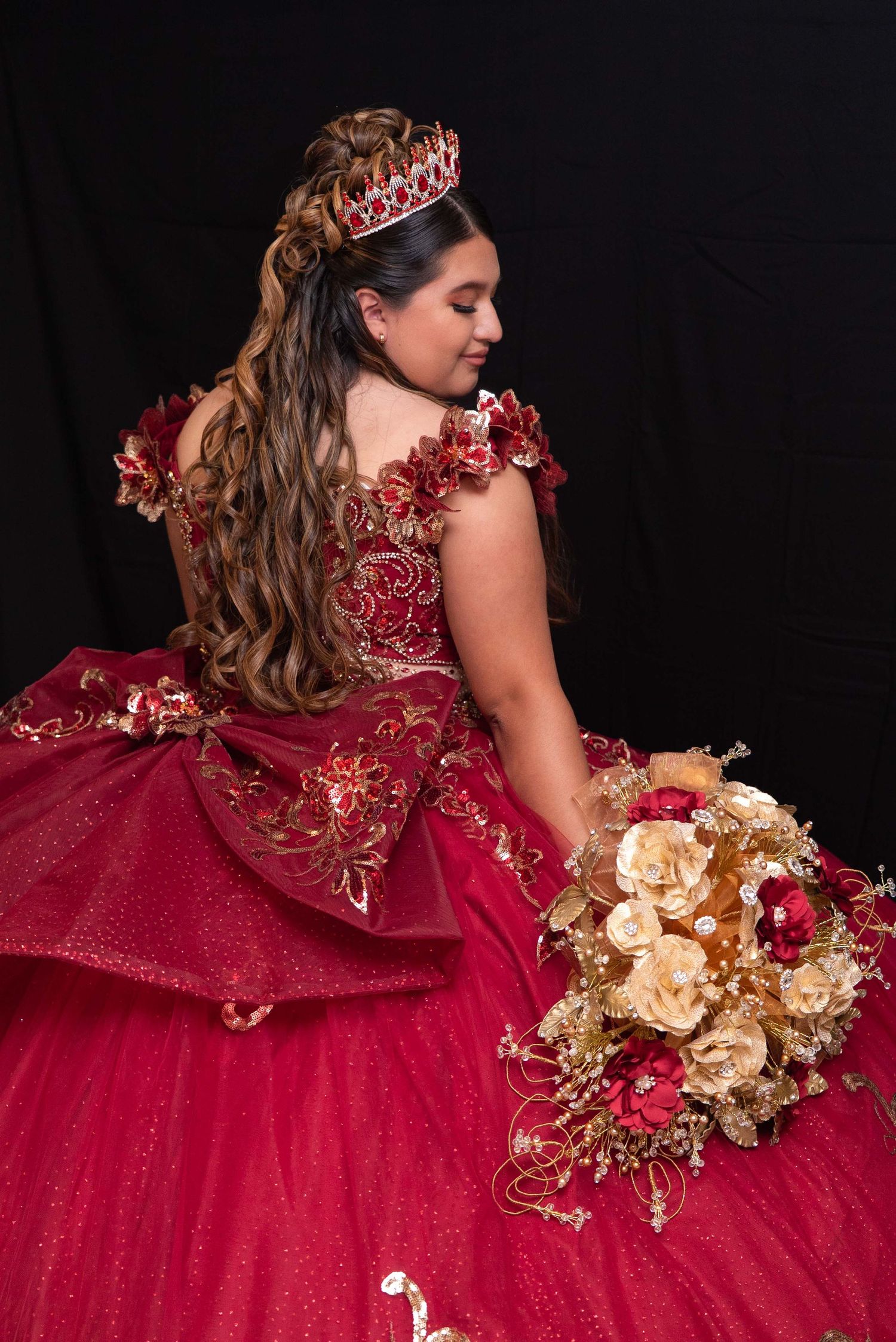 Teen girl in red quinceañera gown with gold embroidery holding bouquet against black backdrop.