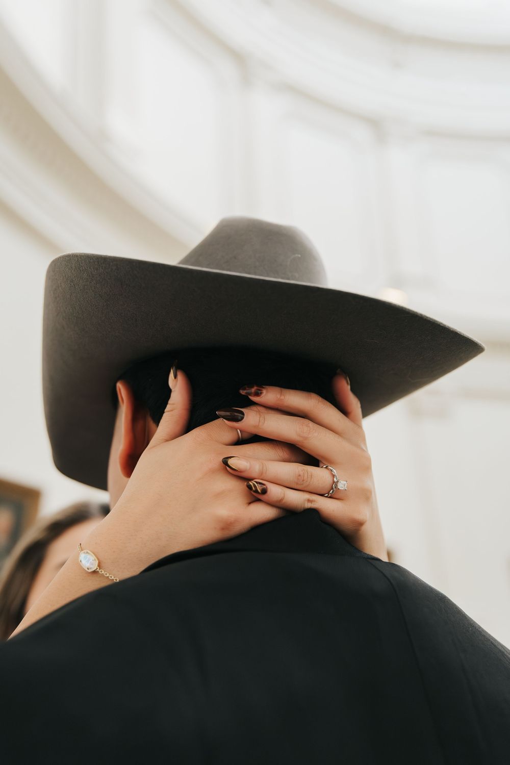 Artistic close-up series showing hands with rings embracing around a black cowboy hat against a white background.