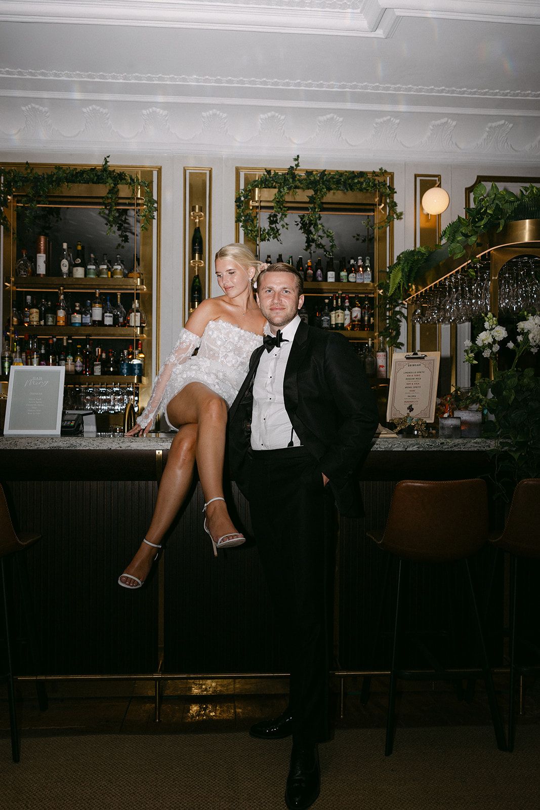 A couple performs a playful dance lift at a bar with bottles and decor visible in the background.