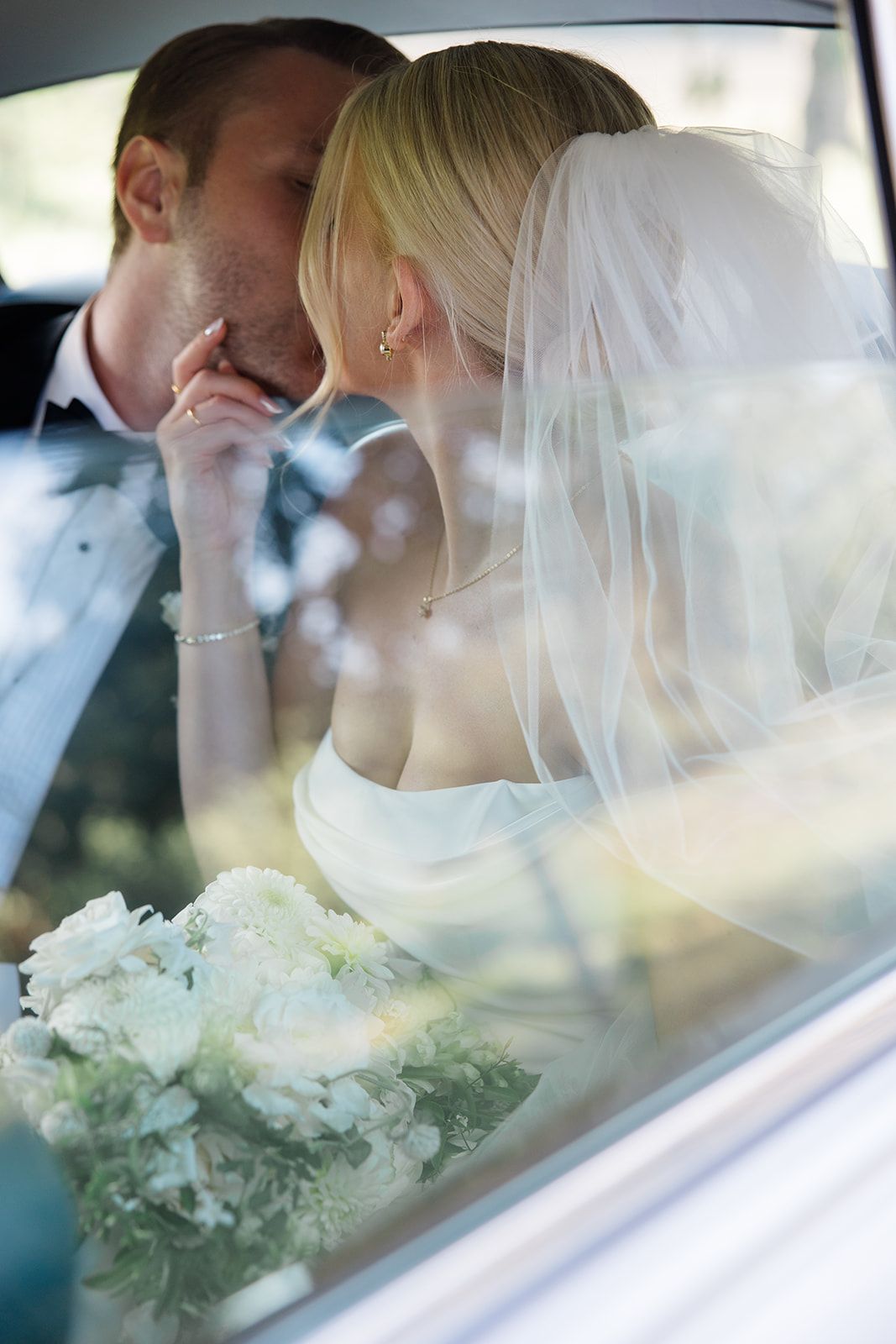 Romantic wedding couple sharing intimate moments through car window with white veil and bouquet visible.