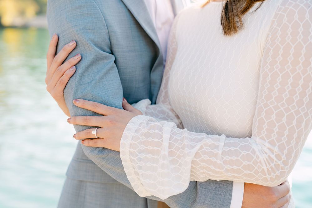 A couple in formal attire embraces by a waterfront setting during a romantic photoshoot at sunset.