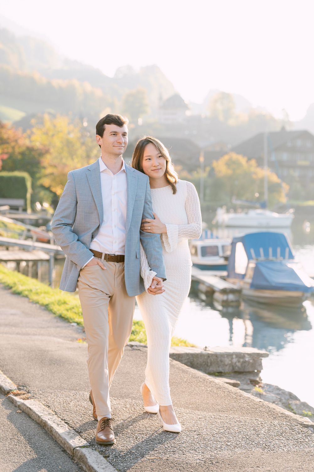 A couple takes a romantic stroll along a marina walkway at sunset with boats in the background.