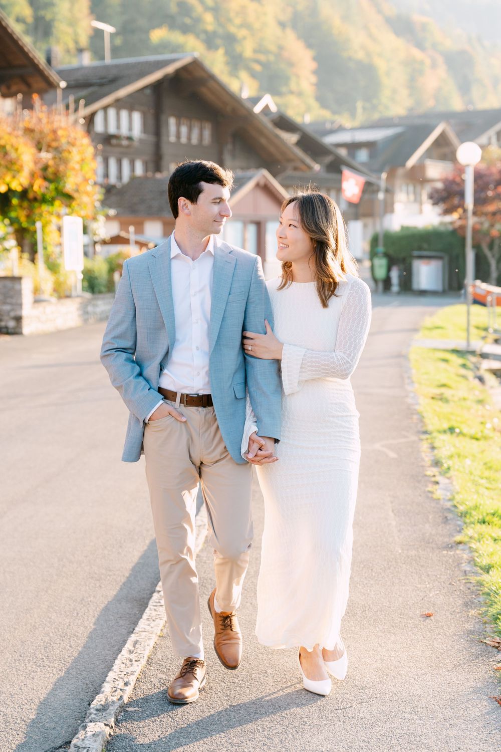 A couple in formal attire walks arm in arm down a street in a charming Swiss village during autumn in Iseltwald.