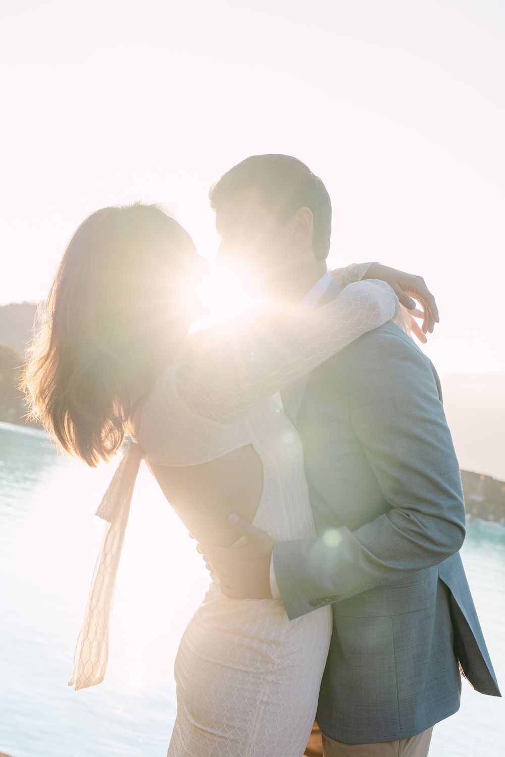 A romantic couple embraces in a dreamy backlit sunset scene by the water creating a soft silhouette effect.