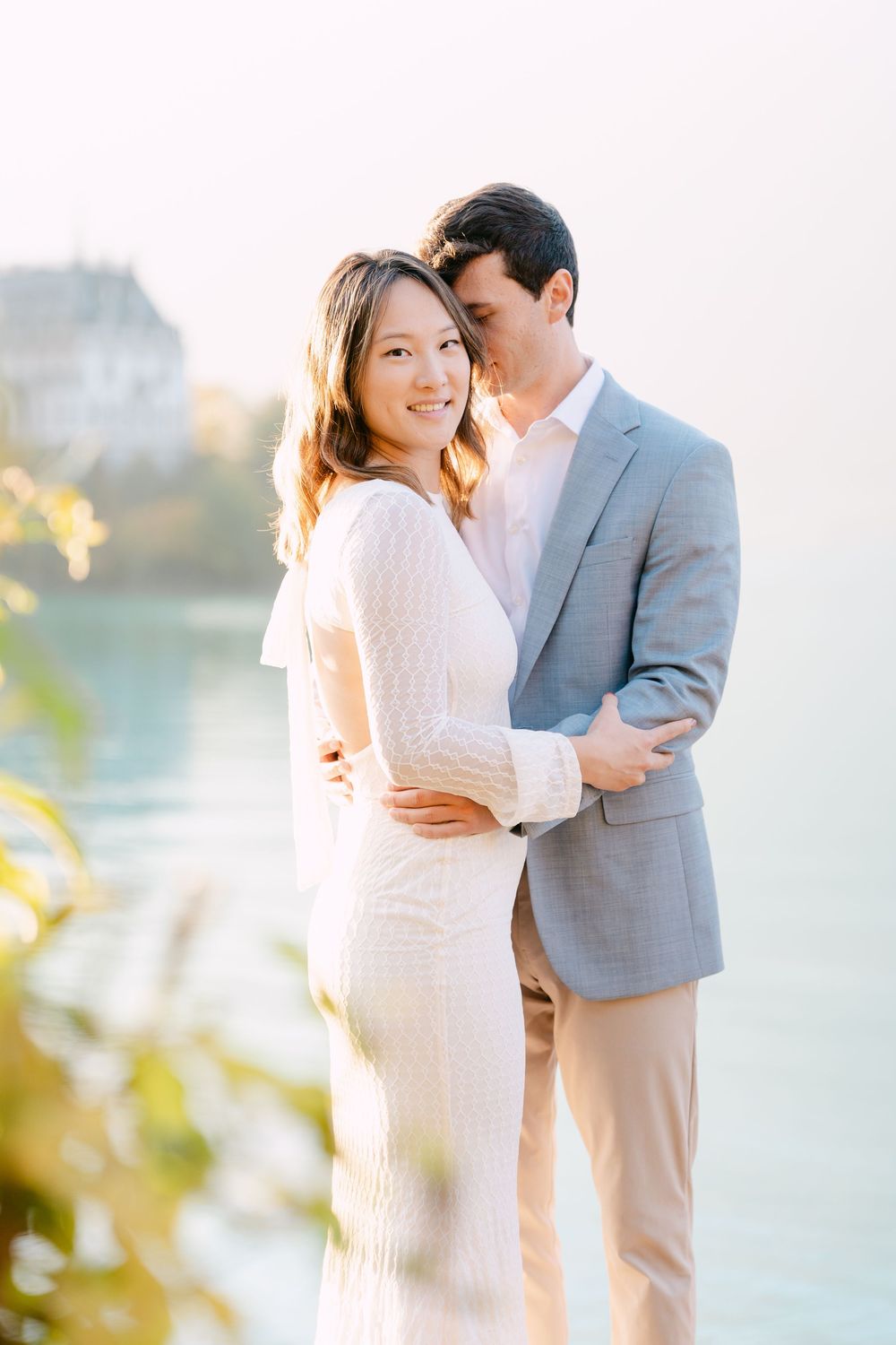 Romantic couple embraces outdoors in elegant attire with soft sunlight and water in the background.