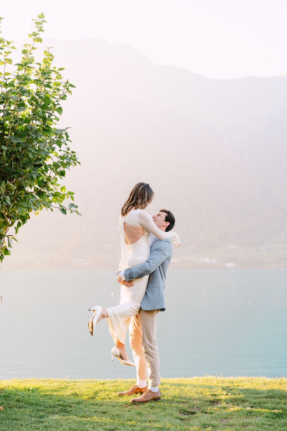 A couple embraces on a grassy hilltop overlooking a serene lake with mountains in the background at sunset.