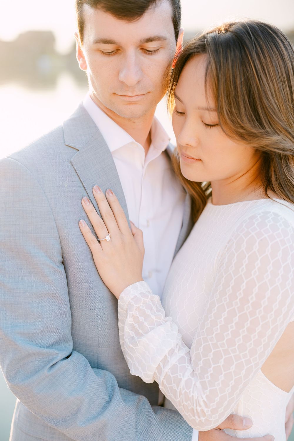 A romantic couple in soft light embrace wearing a grey suit and white lace dress during an intimate photoshoot.