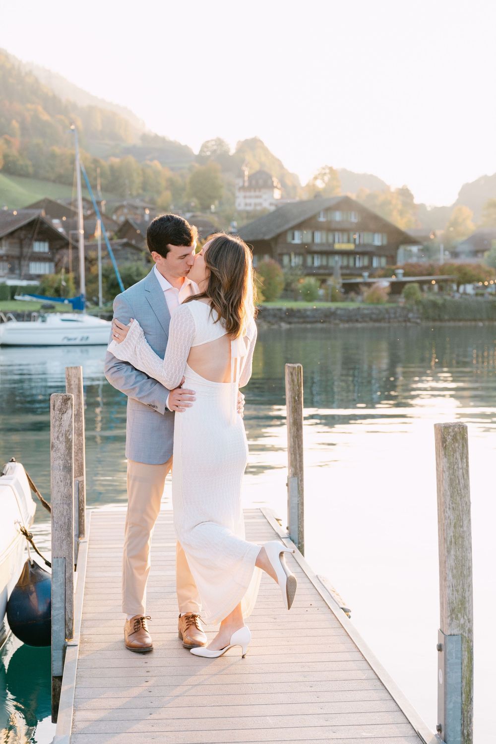 A romantic couple embraces on a wooden dock by a serene lake during golden hour with lakeside homes in the background at the Iseltwald pier.