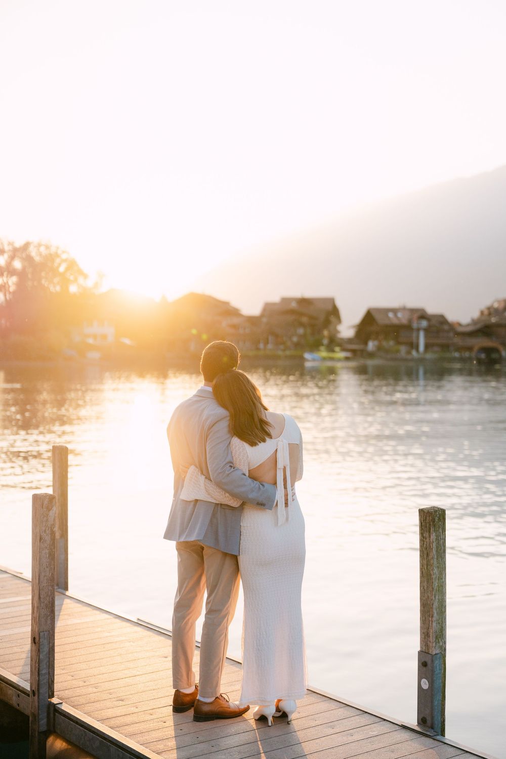 Couple embraces on wooden dock at sunset overlooking tranquil waterfront with houses in background in Iseltwald.