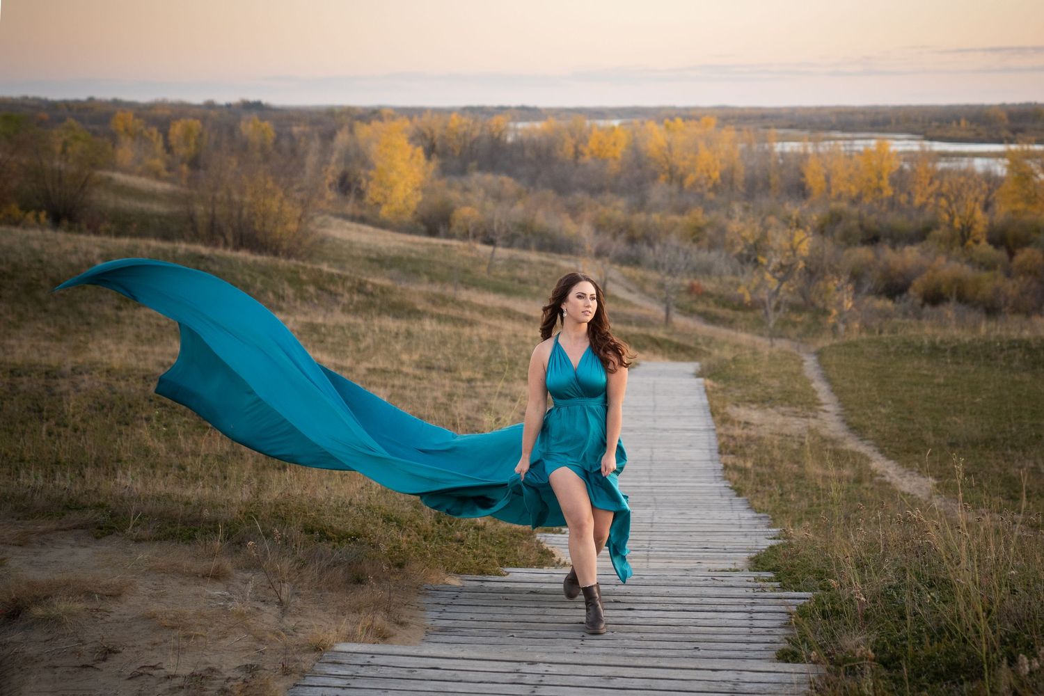 A flowing blue cape billows in the wind on a hilltop overlooking an autumn lake landscape at sunset.