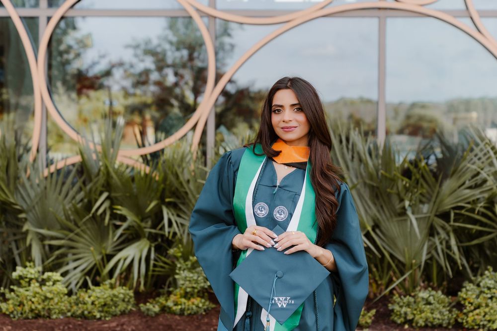 Graduate wearing green academic regalia poses in front of decorative garden arch and tropical plants.