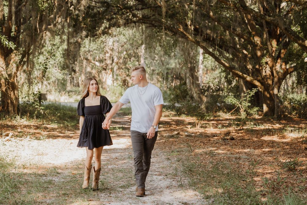 A couple walks hand in hand down a sunlit path through a forest of tall trees.