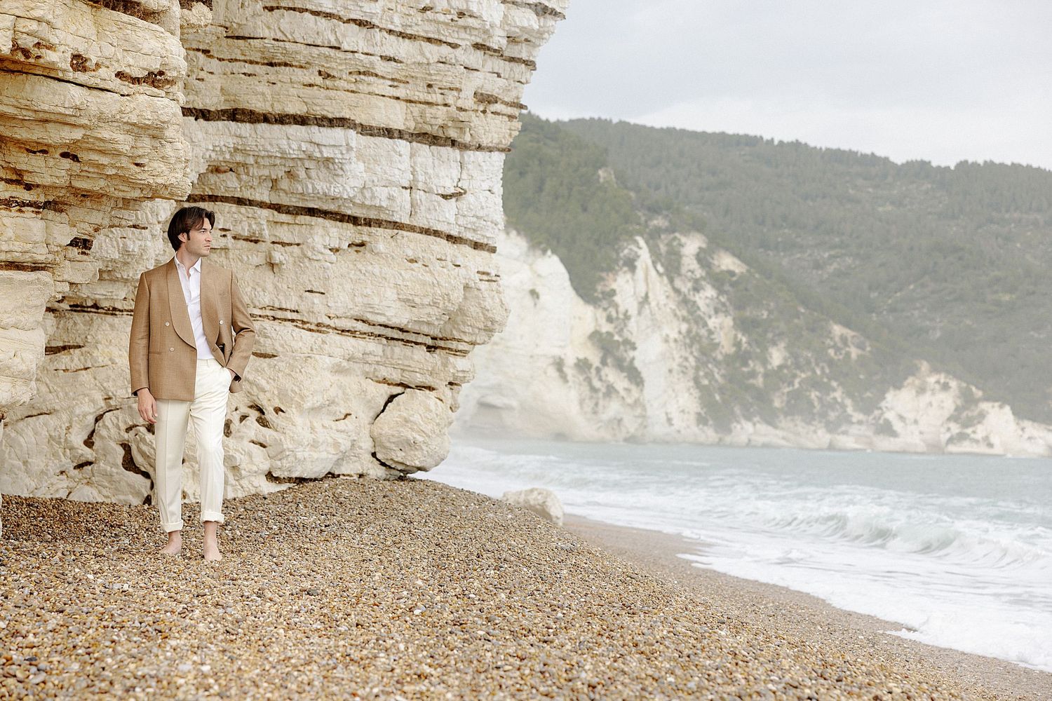 A romantic couple walks along a scenic rocky beach with white cliffs and crashing waves in the background.