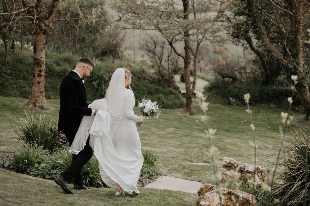 Bride and groom walking through a lush garden, bride holding a white bouquet.