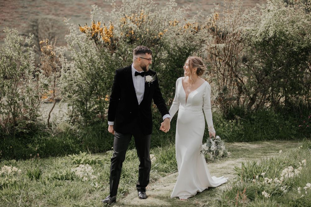 Bride and groom holding hands walking through wildflowers, groom in black tuxedo.