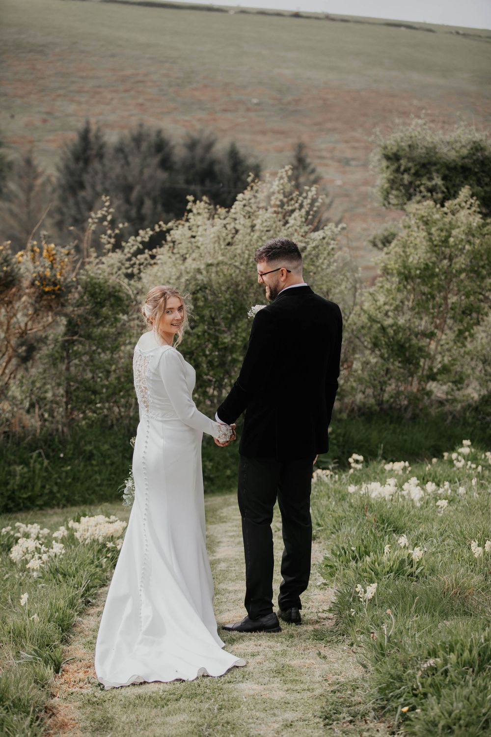Bride in white long-sleeve gown holding groom's hand on a scenic countryside path.