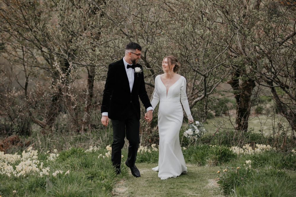 Bride and groom walking hand in hand through a spring orchard with white flowers.