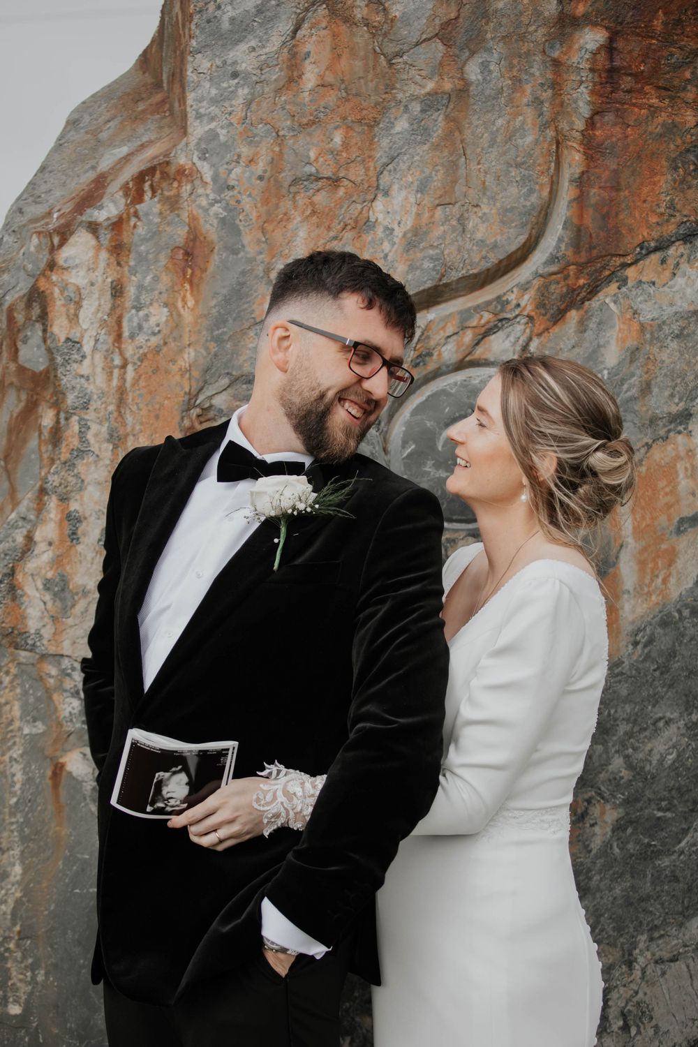 Bride and groom smiling at each other holding ultrasound scan in front of rocky backdrop.
