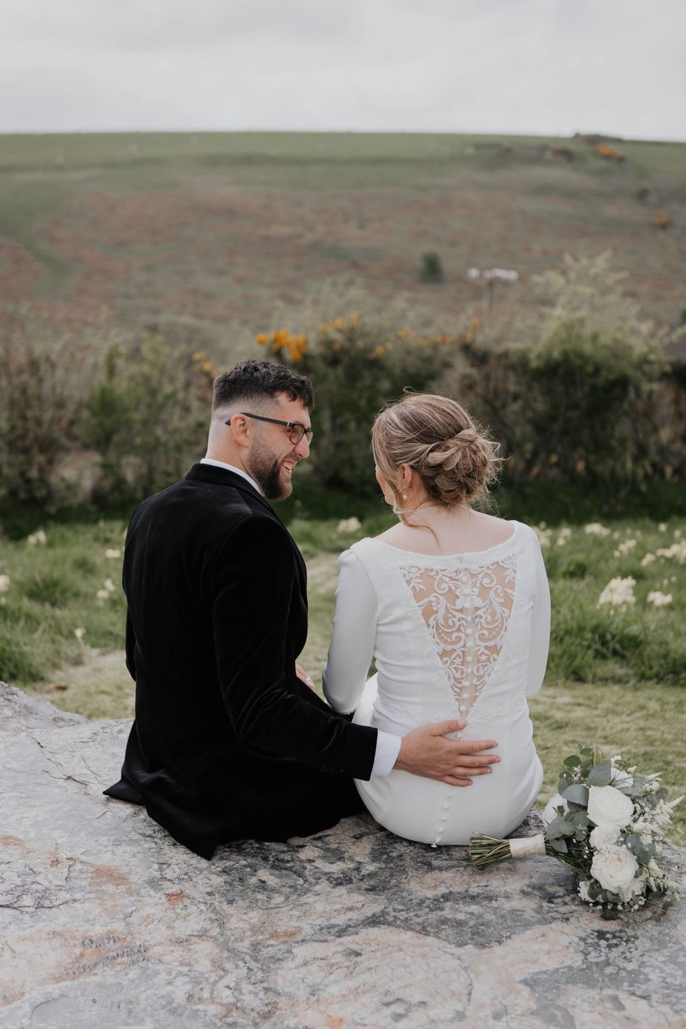Bride and groom sitting on a rock overlooking scenic green rolling hills together.