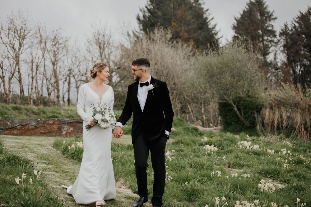 Bride and groom walking hand in hand through a garden path lined with white flowers.