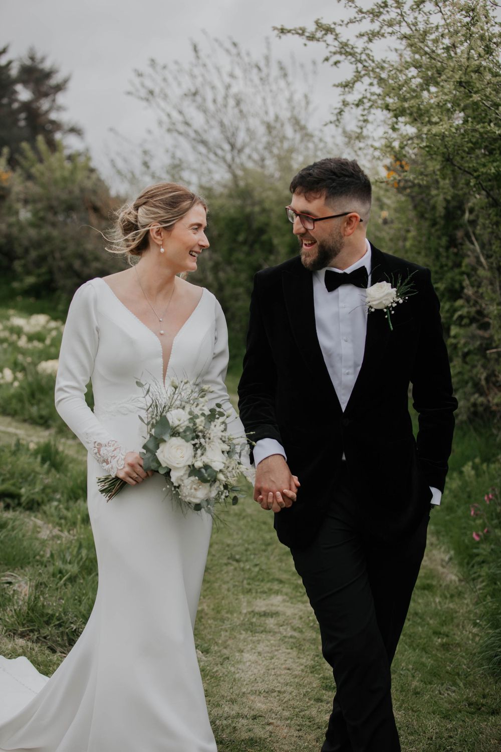 Smiling bride and groom walking together outdoors, bride holding white rose bouquet.