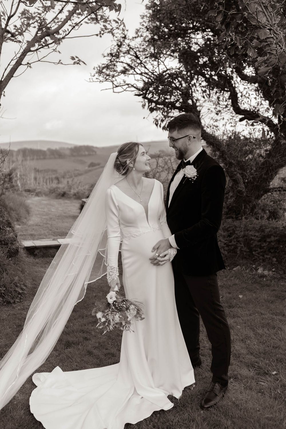 Black and white photo of bride in flowing veil and groom holding hands outdoors.