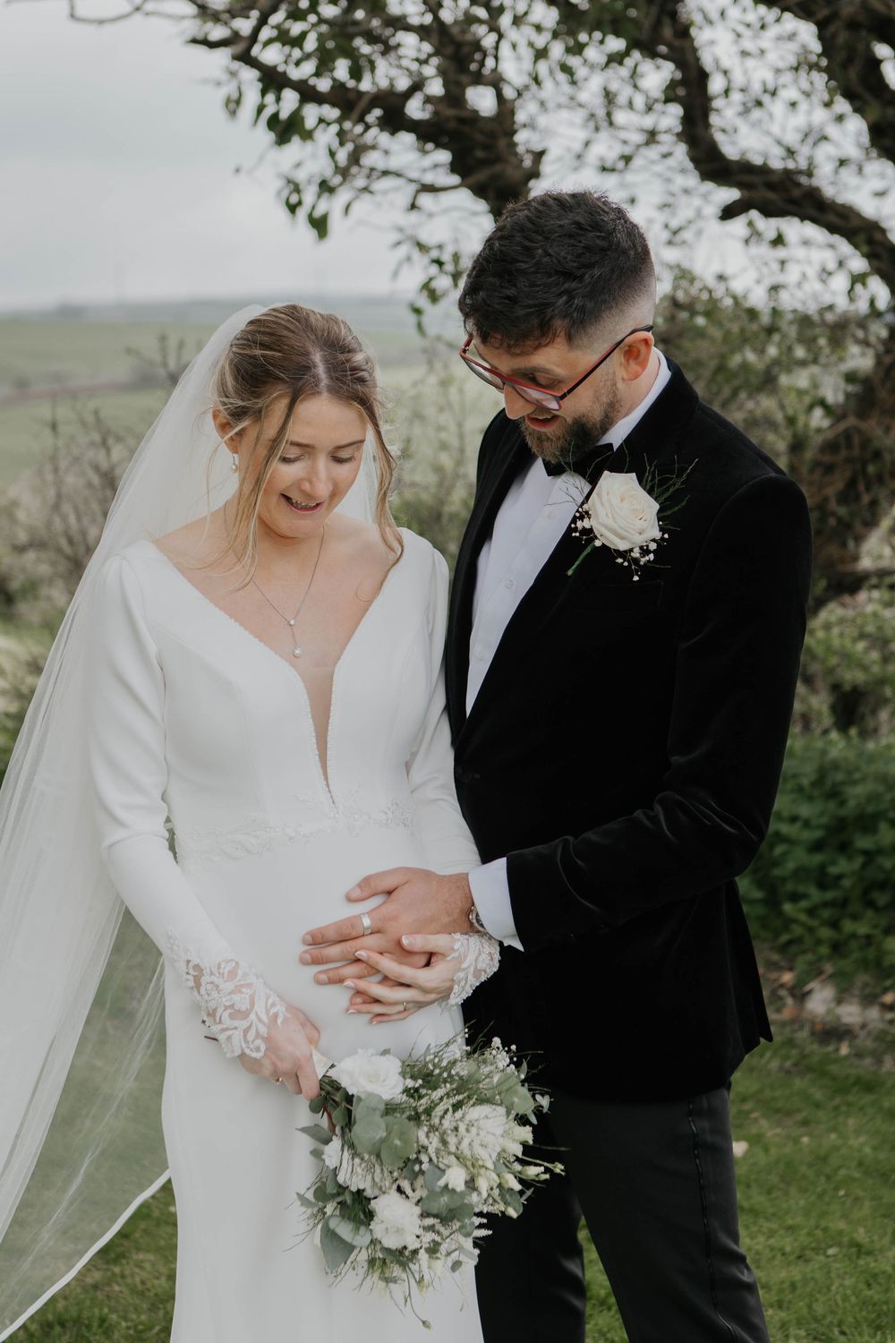 Groom tenderly placing hands on pregnant bride's bump while she holds white bouquet.
