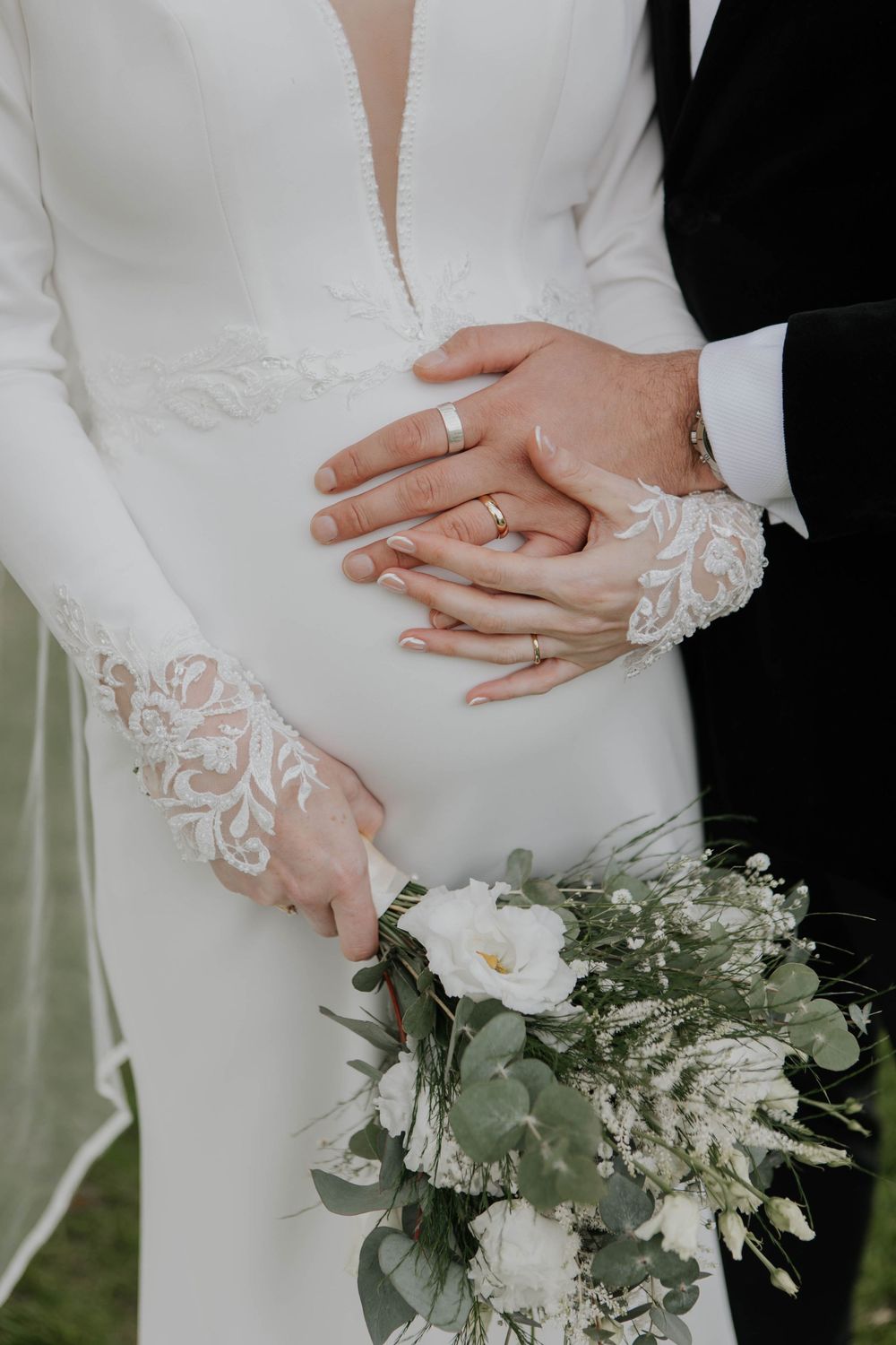 Close-up of bride and groom's hands with wedding rings resting on pregnant belly.