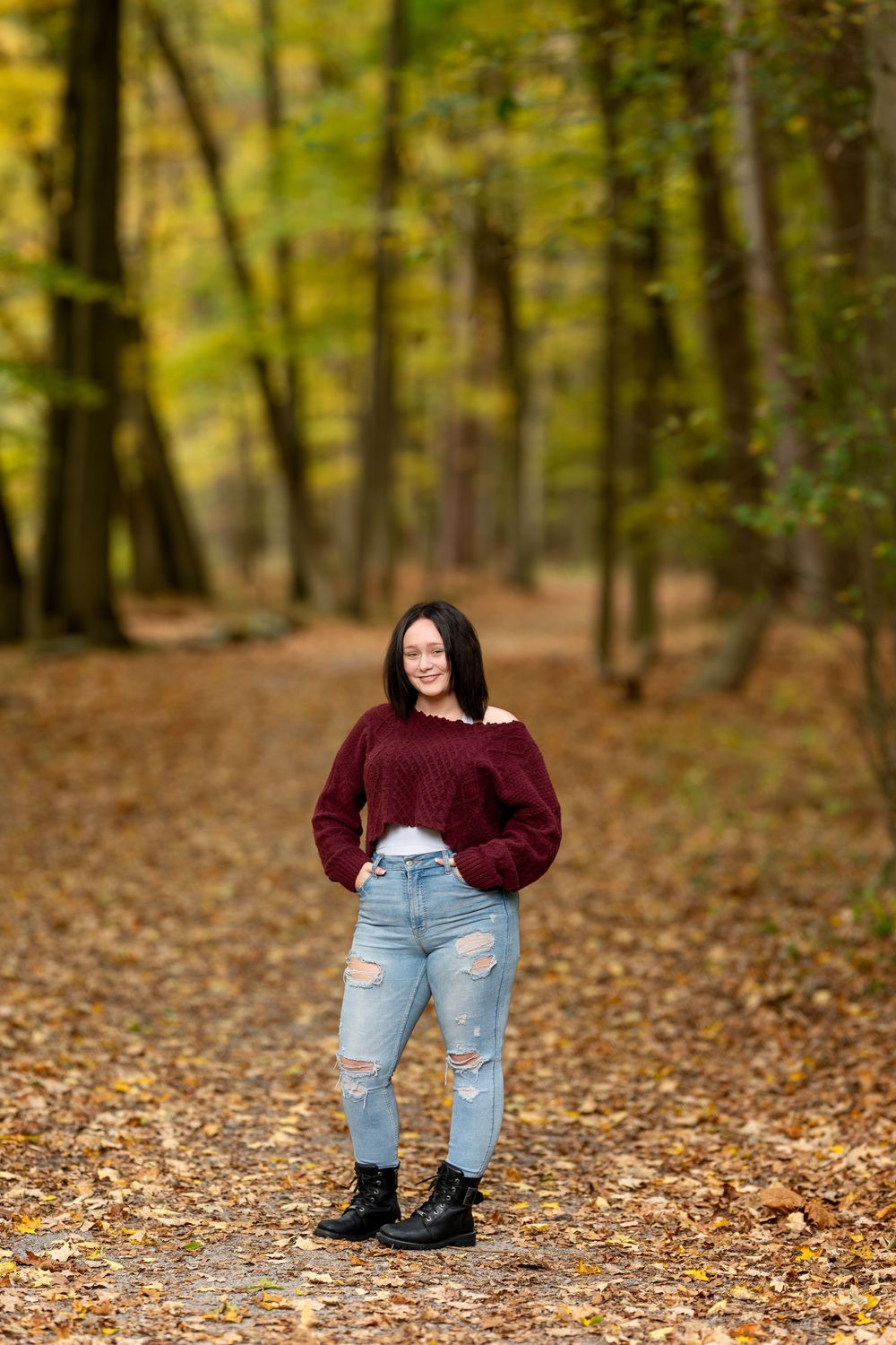 Smiling brunette in burgundy sweater, ripped jeans, and black boots stands on scenic fall forest path.