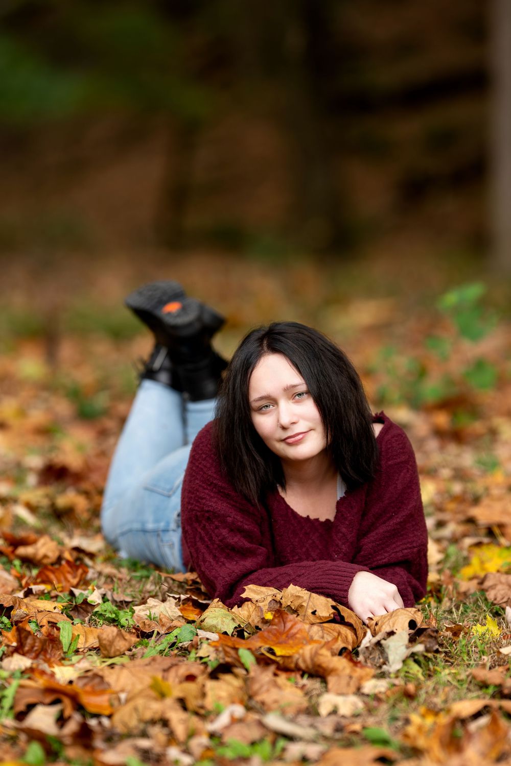 Brunette girl in burgundy knit sweater relaxing on ground covered with colorful fall leaves outdoors.